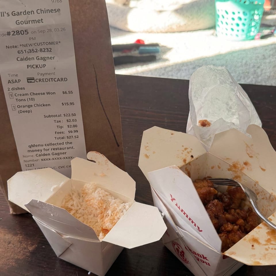 Takeout containers of fried rice and orange chicken sit on a wooden table beside a Bill’s Garden Chinese restaurant receipt.